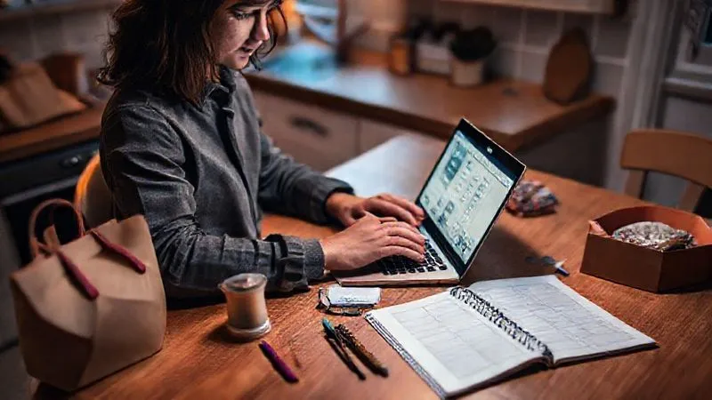 A photorealistic image of a person working diligently on a laptop at a kitchen table, surrounded by subtle visual cues of various side hustles: a small delivery bag, a craft project, and a planner open to a freelance schedule. The scene is lit with warm, ambient light, conveying productivity and multi-tasking. 8K, cinematic lighting, sharp focus, depth of field, shot on a high-end DSLR.
